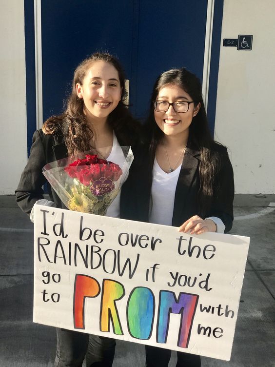 Two girls holding flowers and a sign that says "I'd be over the rainbow if you'd go to prom with me."